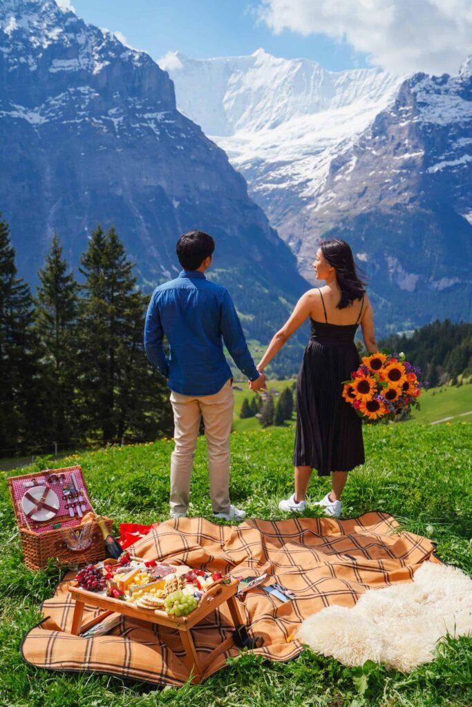 Couple holding hands, facing Swiss mountain view with picnic setup and bouquet in foreground