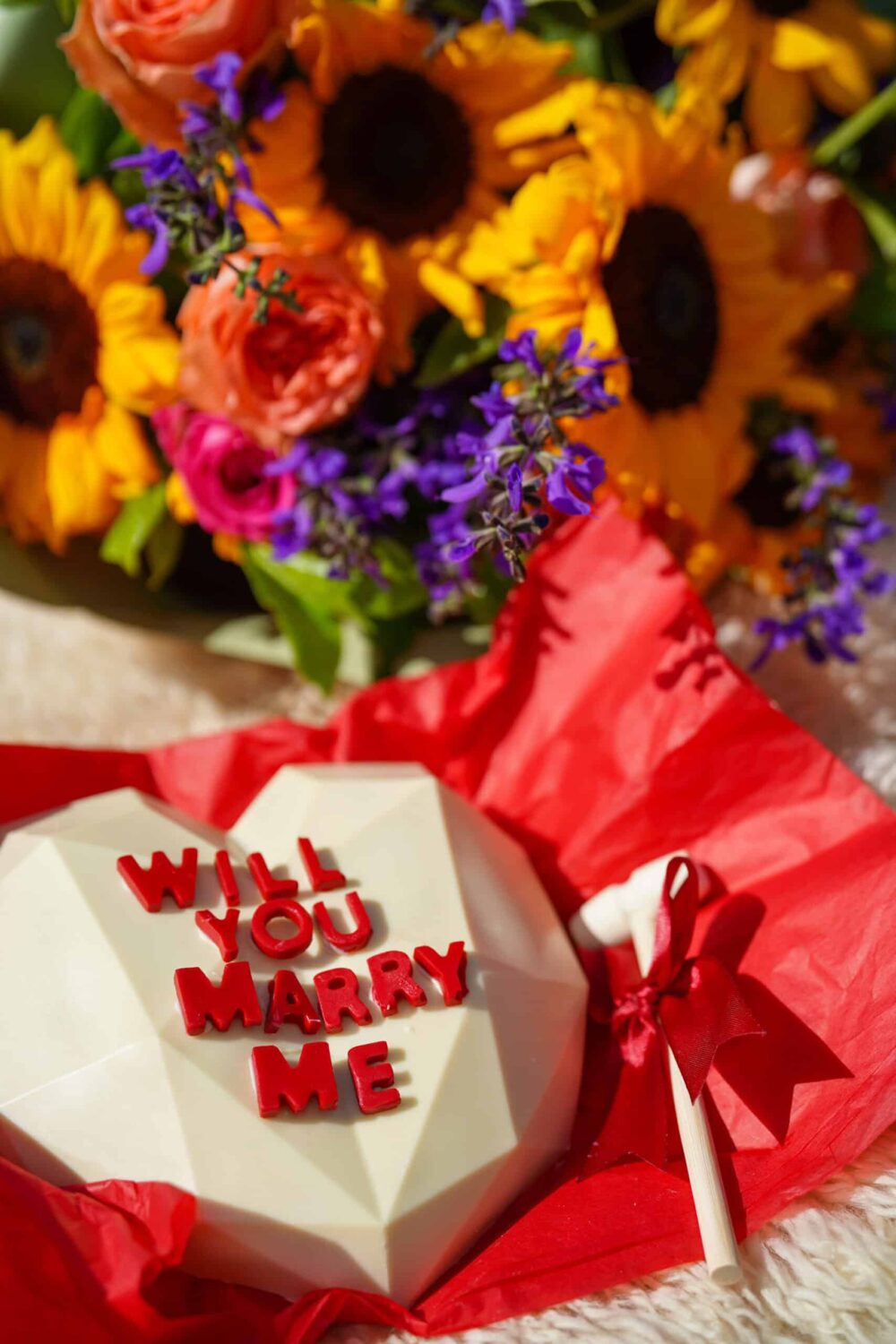 Close-up of colorful bouquet with “Will You Marry Me” chocolate heart, part of picnic setup.