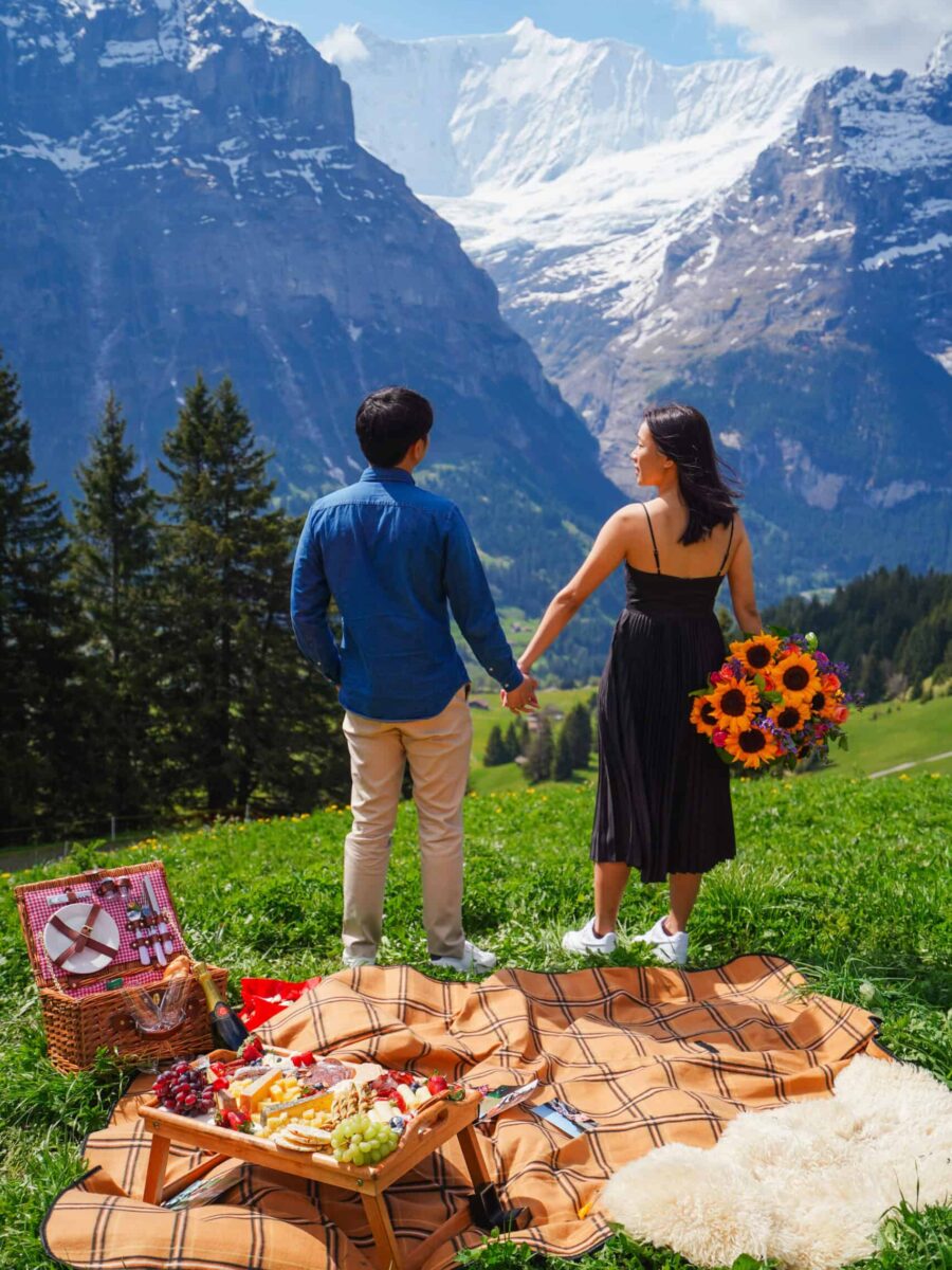 Couple holding hands, facing Swiss mountain view with picnic setup and bouquet in foreground